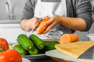 A woman in an apron on the background of the kitchen peels carrots from the skin