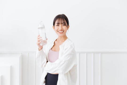 Asian woman with long black hair tied up, wearing sportswear and a white sports jacket. Happy smiling at the camera and holding a water bottle standing on a white background. Image with copy space.