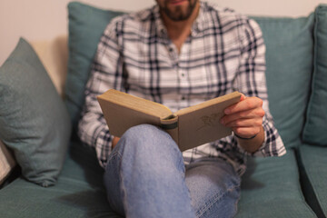 Closeup view of male hands holding book. Young man relaxing at home sitting in a couch. Selective focus on hand, blurred background. Horizontal.