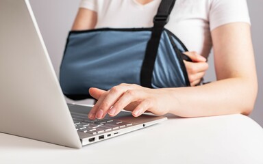 Woman with painful arm in sling working on laptop. Female wearing bandage to support injured elbow,...