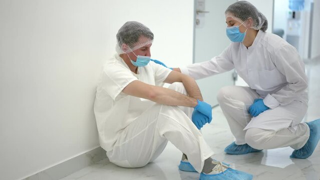 Depressed Caucasian Man Sitting On Floor In Hospital Corridor Leaning On Wall As Colleague Touching Shoulder Endorsing Coworker. Portrait Of Desperate Exhausted Sad Doctor Supported By Nurse In Clinic