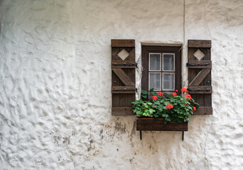 A small wooden window with window shutters and a flower pot with red flowers