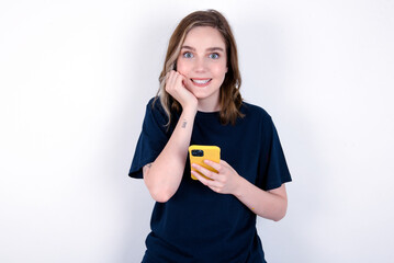 Fototapeta na wymiar Joyous young caucasian woman wearing black T-shirt poses with mobile phone device, types text message on modern smartphone, watches funny video during free time, enjoys good internet connection,