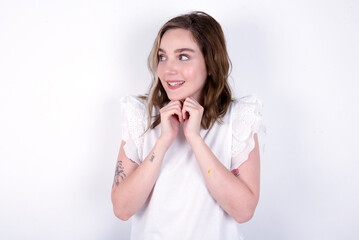 Happy young caucasian woman wearing white T-shirt over white background anticipates something awesome happen, looks happily aside, keeps hands together near face, has glad expression.