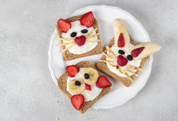 Three sweet toasts for kids in shape of cat, rabbit and chick with strawberries, banana, cream cheese and coconut flakes on round plate on gray background