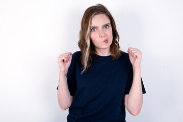 Irritated young caucasian woman wearing black T-shirt over white background blows cheeks with anger and raises clenched fists expresses rage and aggressive emotions. Furious model