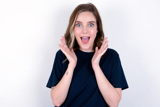Surprised happy young caucasian woman wearing black T-shirt over white background , glad to see big discounts on clothes, expresses shock, keeps hands near head, jaw dropped.