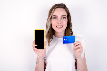 Photo of adorable young caucasian woman wearing white T-shirt over white background holding credit card and Smartphone. Reserved for online purchases