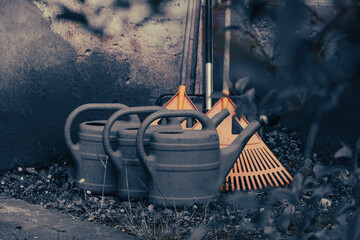 Close-up of plastic watering cans and leaf rakes placed against a concrete wall.