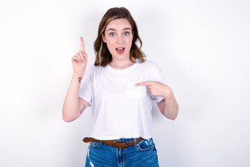 young caucasian woman wearing white T-shirt over white background says: wow how exciting it is, has amazed expression, indicates something. One hand on her chest and pointing with other hand.