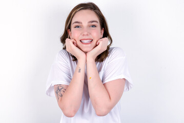 Fototapeta premium Dreamy young caucasian woman wearing white T-shirt over white background keeps hands pressed together under chin, looks with happy expression, has toothy smile.