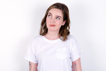 young caucasian woman wearing white T-shirt over white background has worried face looking up lips together, being upset thinking about something important, keeps hands down.