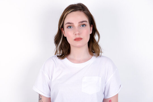 Joyful young caucasian woman wearing white T-shirt over white background looking to the camera, thinking about something. Both arms down, neutral facial expression.