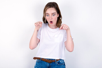 Amazed young caucasian woman wearing white T-shirt over white background points down with fore...