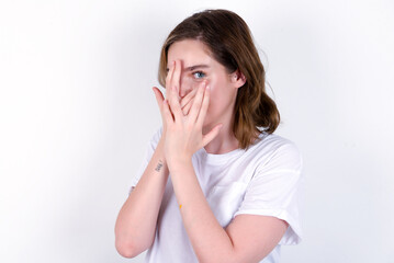 young caucasian woman wearing white T-shirt over white background covering face with hands and peering out with one eye between fingers. Scared from something or someone.