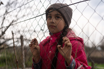 A little girl with a sad look behind a metal fence. Social problem of refugees and forced migrants