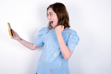 Happy cheerful young caucasian woman wearing blue T-shirt over white background receiving good news via e-mail and celebrating success while standing and looking at mobile phone.