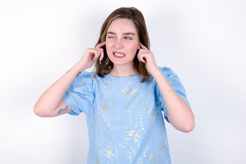 Happy young caucasian woman wearing blue T-shirt over white background ignores loud music and plugs ears with fingers asks to turn off sound