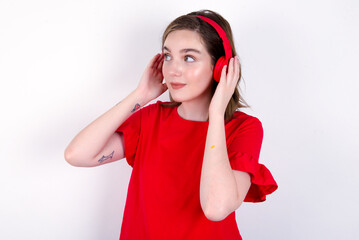 Obraz na płótnie Canvas young caucasian woman wearing red T-shirt over white background wears stereo headphones listens music concentrated aside. People hobby lifestyle concept
