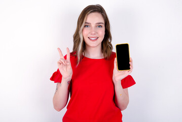 Naklejka premium young caucasian woman wearing red T-shirt over white background holding modern device showing v-sign