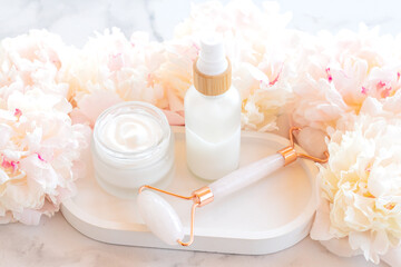 Facial kit for home skincare and spa. Rose quartz Face roller and jar of face cream on white marble background with peony flowers. Natural treatment concept.