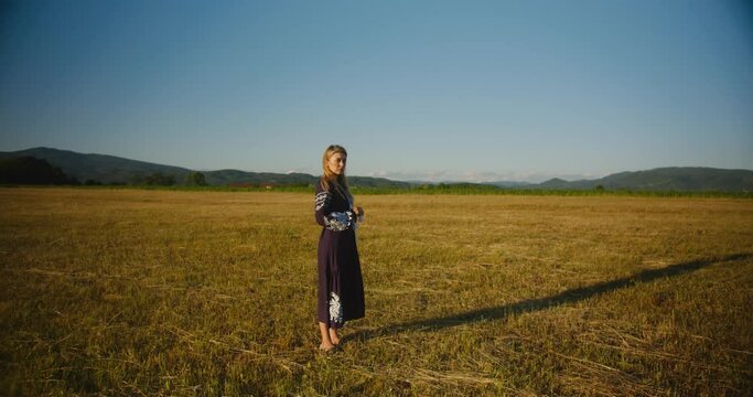 A romantic girl in a national ukrainian dress stands in a wheat field in the rays of the evening setting sun or rising sun, playing with wheat ears, acting on camera