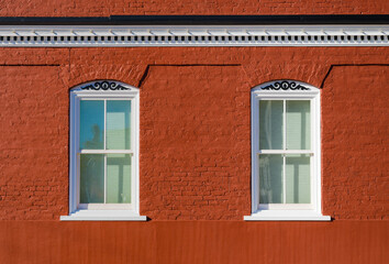 Two white framed windows in a rich colored red brick building with scrollwork and cornice with a...