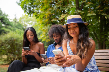 Three friends sitting on a bench in a park use their smartphones, multi-ethnic group