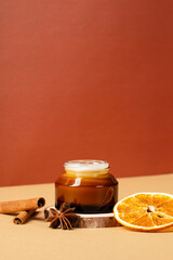 Amber glass jar of moisturizer cream with wooden branch, slice of dry orange, cinnamon sticks....
