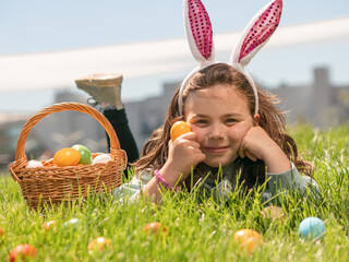 A little girl in rabbit ears is lying on the green grass with a basket full of Easter eggs, holding...