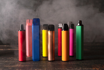 Set of colorful disposable electronic cigarettes on a dark wood background with smoke. - obrazy, fototapety, plakaty