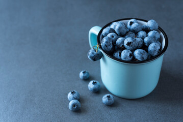 Ripe blueberry in a blue mug and a few berries nearby on the dark grey background. Top view. Copy space. Minimalism. Healthy nutrition