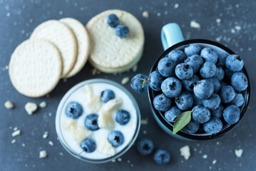 Composition of ripe blueberry in a mug, yogurt in a glass bowl, cookies and scattered crumbs around on the dark grey background. Flat lay. Top view. Healthy nutrition