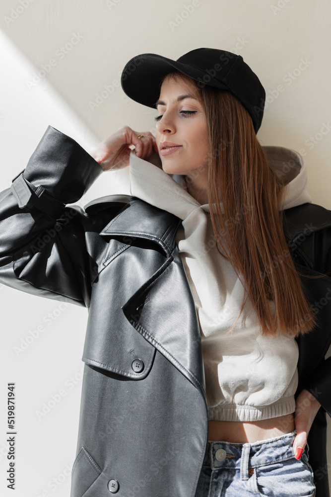 Sticker Beautiful trendy young hipster woman with a fashion black mockup cap in a stylish leather coat with a fashionable hoodie stands and poses near a beige wall in the sunlight. Urban female style look