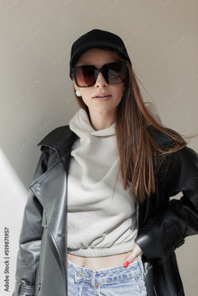 Canvas Prints Stylish beautiful hipster girl with fancy vintage sunglasses and fashion black cap in a black leather jacket and hoodie sweatshirt stands near the wall in the sunlight