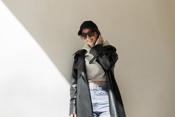 Trendy fashionable happy beautiful young girl with a smile with cool sunglasses and a black cap mockup in a fashion leather coat with a hoodie and jeans stands near the wall in the street with light