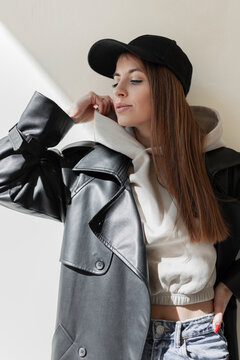 Beautiful trendy young hipster woman with a fashion black mockup cap in a stylish leather coat with a fashionable hoodie stands and poses near a beige wall in the sunlight. Urban female style look