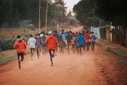 Group Of Runners On A Morning Mass Training In Kenya. Africans Running On Red Roads In Iten City. Running Preparation For Marathon Races