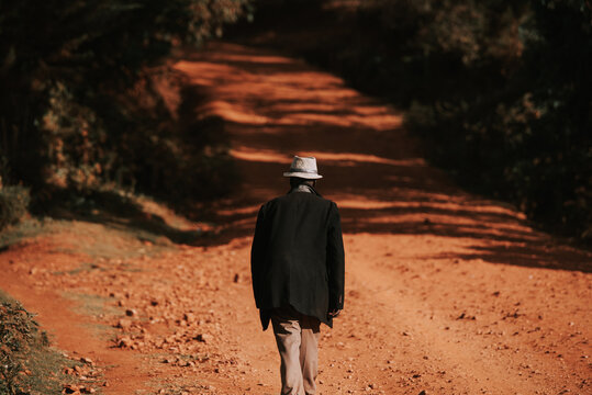 An Old Man Walks Along The African Red Roads. Illustrative Photo Of Life In Africa And Kenya. Life Of A Retired Pensioner In East Africa