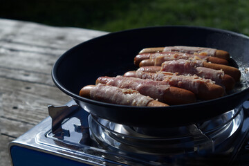 Sausages wrapped in bacon are fried in a frying pan on a gas camping stove against the backdrop of nature