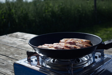Sausages wrapped in bacon are fried in a frying pan on a gas camping stove against the backdrop of...