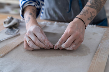 Woman potter standing at work table working with clay at workshop, cropped photo. Close up of tattooed female hands making handmade pottery, molding shaping ceramic plate in creative handcraft studio