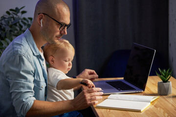Fototapeta premium Young father working remotely on laptop with little baby son in his arms
