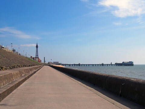 vertical perspective view along the pedestrian promenade in blackpool with a view of the town tower and pier in the distance with calm summer sea and blue sky