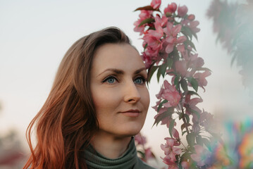 A headshot of a redhead woman is staring into the distance with flowering sakura in the background at sunset.