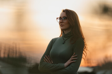 A happy redhead woman in sunglasses with her arms folded across her chest is posing in the background of the cloudy evening sky