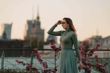 A happy redhead woman in glasses is staring into the distance with warsaw skyscrapers in the background at sunset.