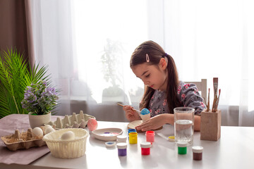 Obraz na płótnie Canvas A cute little girl sits at a white table by the window, paints eggs with a brush and paint in jars.