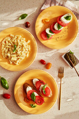 Mediterranean pasta fusilli, Italian caprese salad - tomatoes, mozzarella, basil and pesto. Bread appetizer with caprese salad on orange plates under trendy shadows. Top view food flat lay.