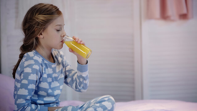 Preteen girl in pajama drinking orange juice on blurred bed in evening.